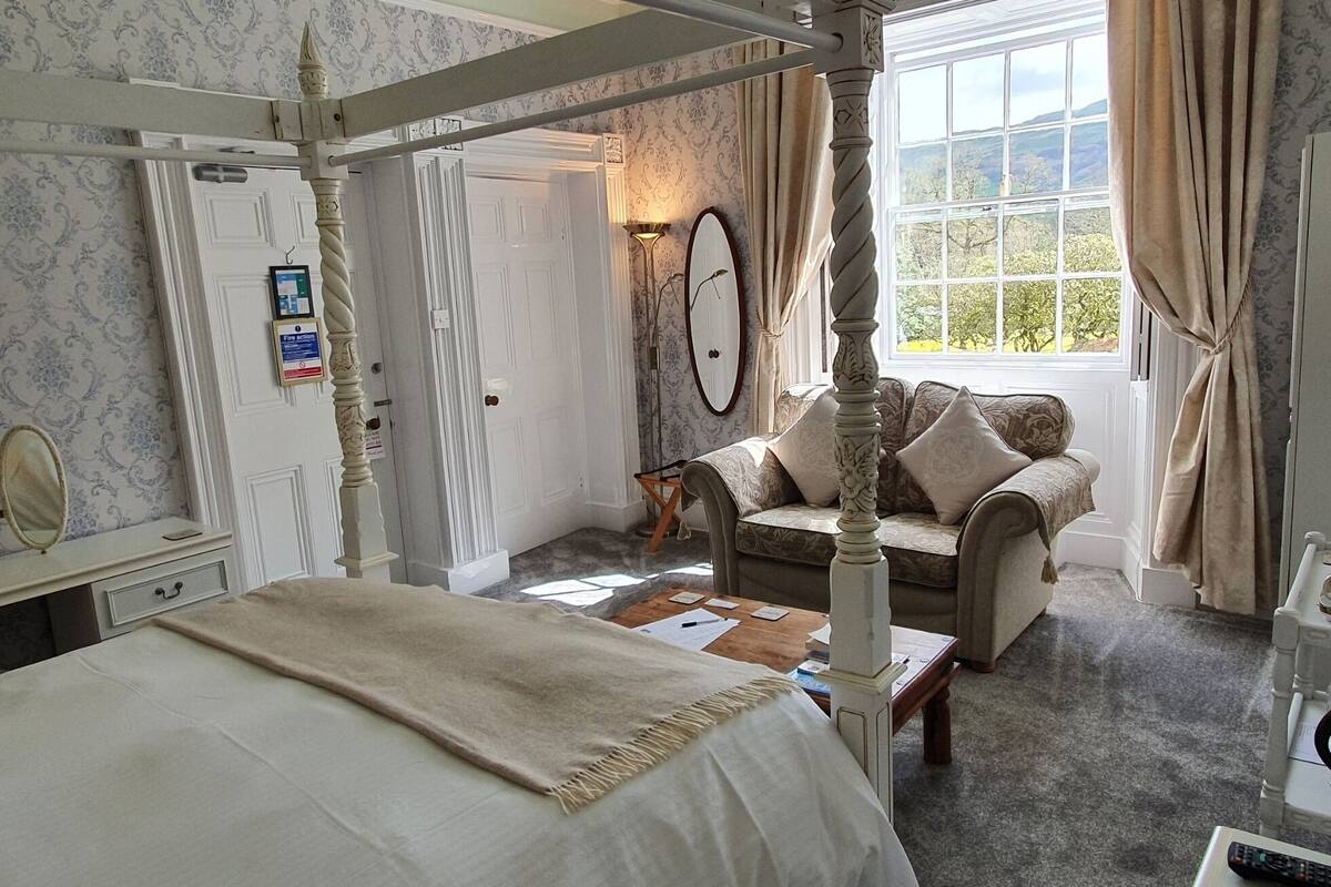 A traditional four-poster bed with a canopy, a sofa, a mirror, and a window with curtains.