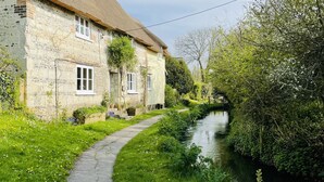 A narrow stone pathway leading to a traditional stone house with a thatched roof, surrounded by lush greenery and a small waterway.