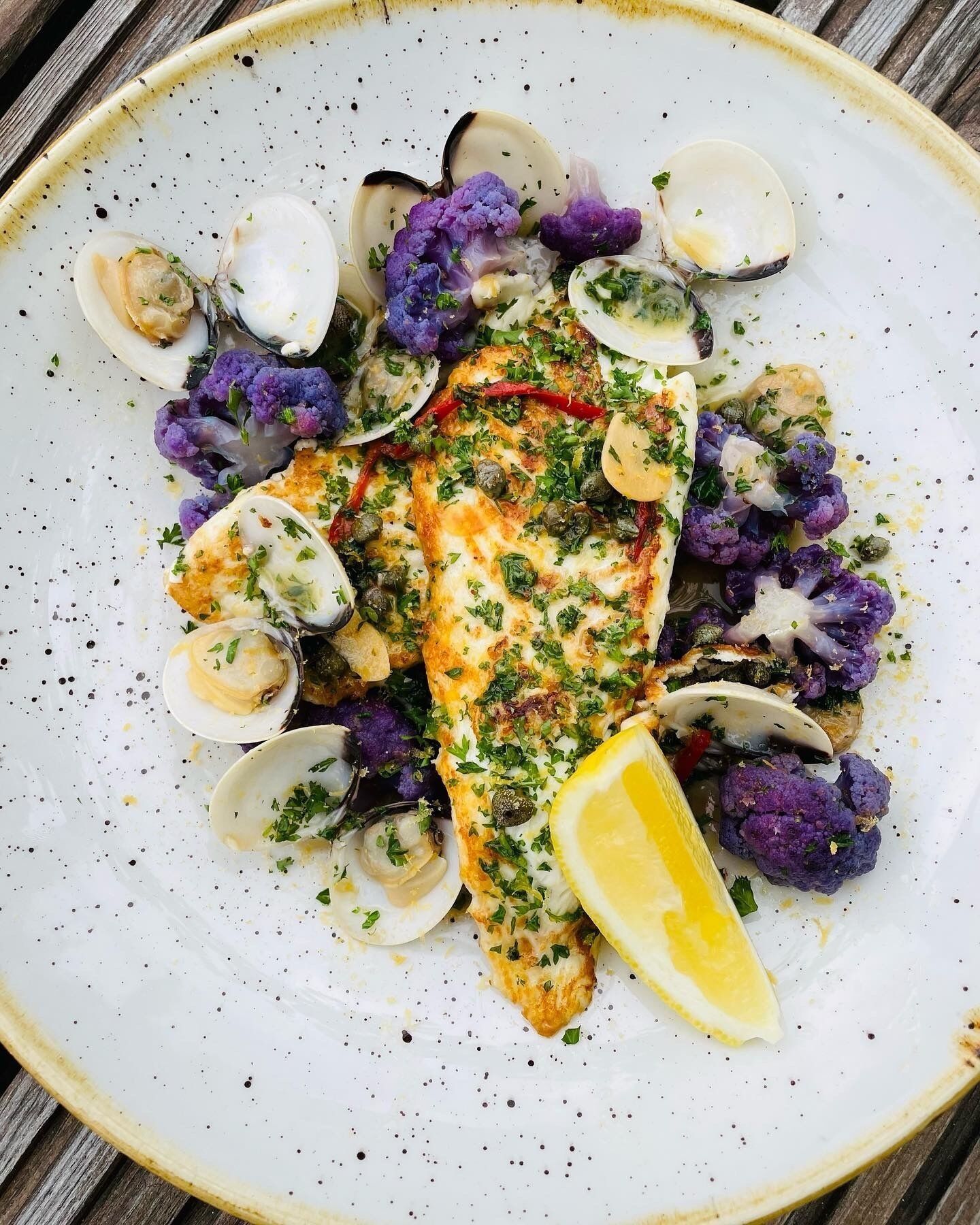 A plate of food with clams, cauliflower, and a piece of fish, garnished with herbs and lemon.