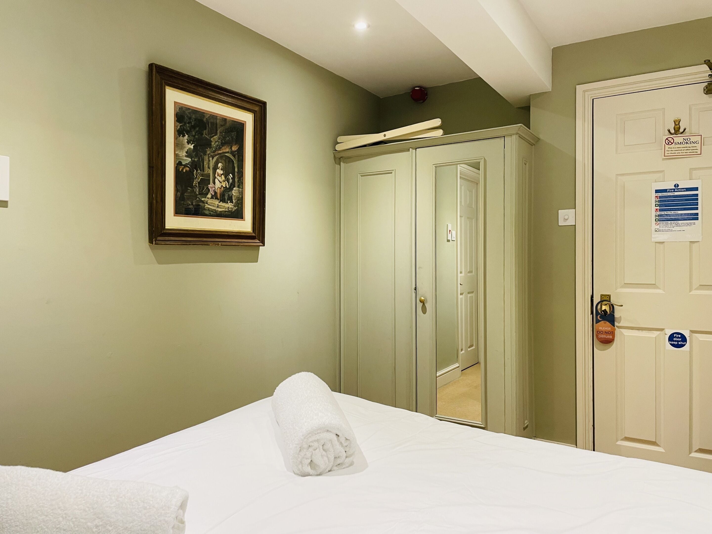 Une chambre d’hôtel avec un lit, un tableau encadré au mur et une porte sur laquelle est apposé un panneau « Il est interdit de fumer ».