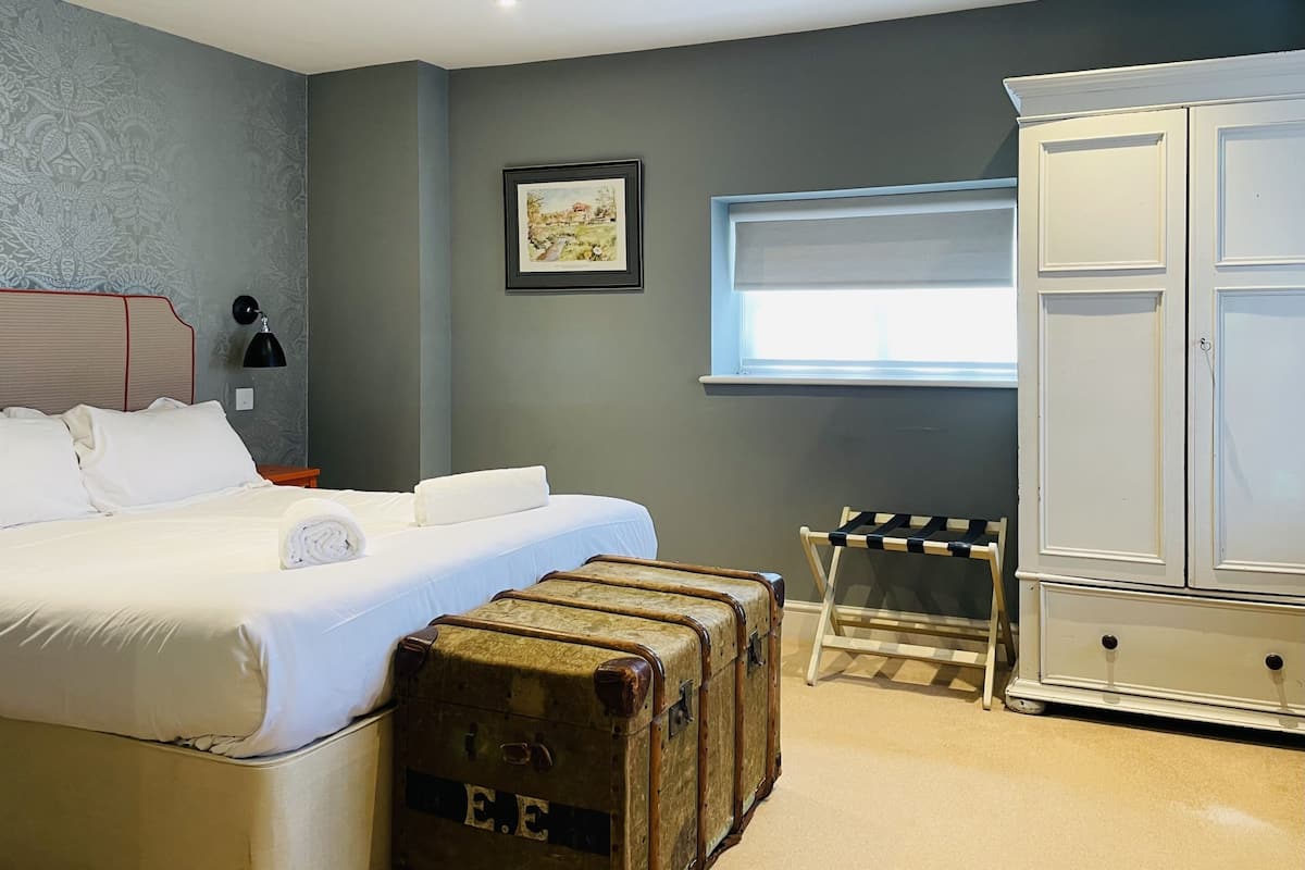 A bedroom with a bed, a nightstand, a lamp, a wardrobe, a window, and a suitcase.