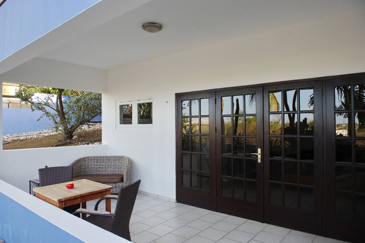 Un balcón con una silla de mimbre, una mesa de madera con un objeto rojo, y vistas a un árbol y la playa.