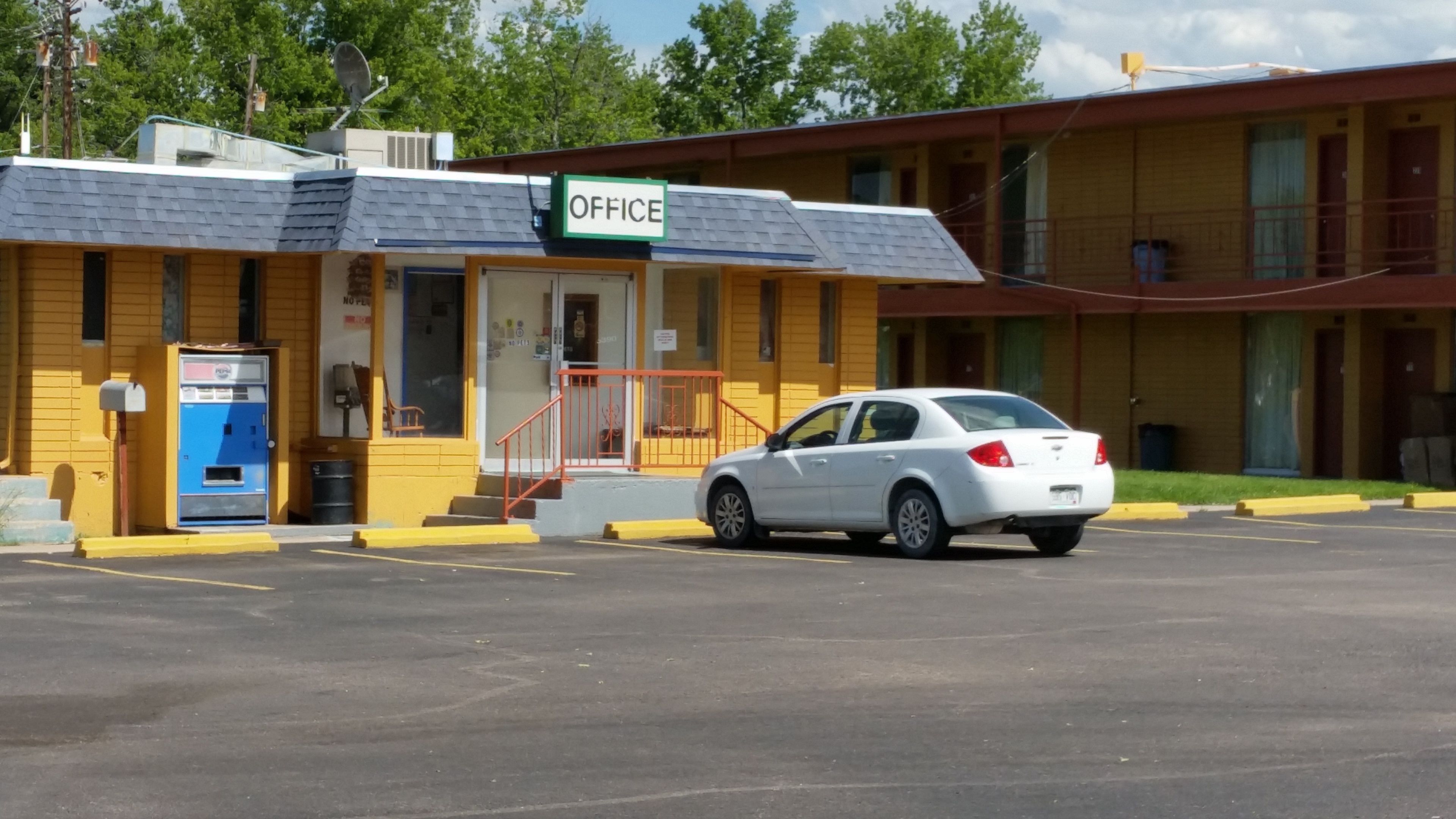 Un motel con un cartello 'OFFICE', una cassetta delle lettere blu e un'auto bianca parcheggiata davanti.