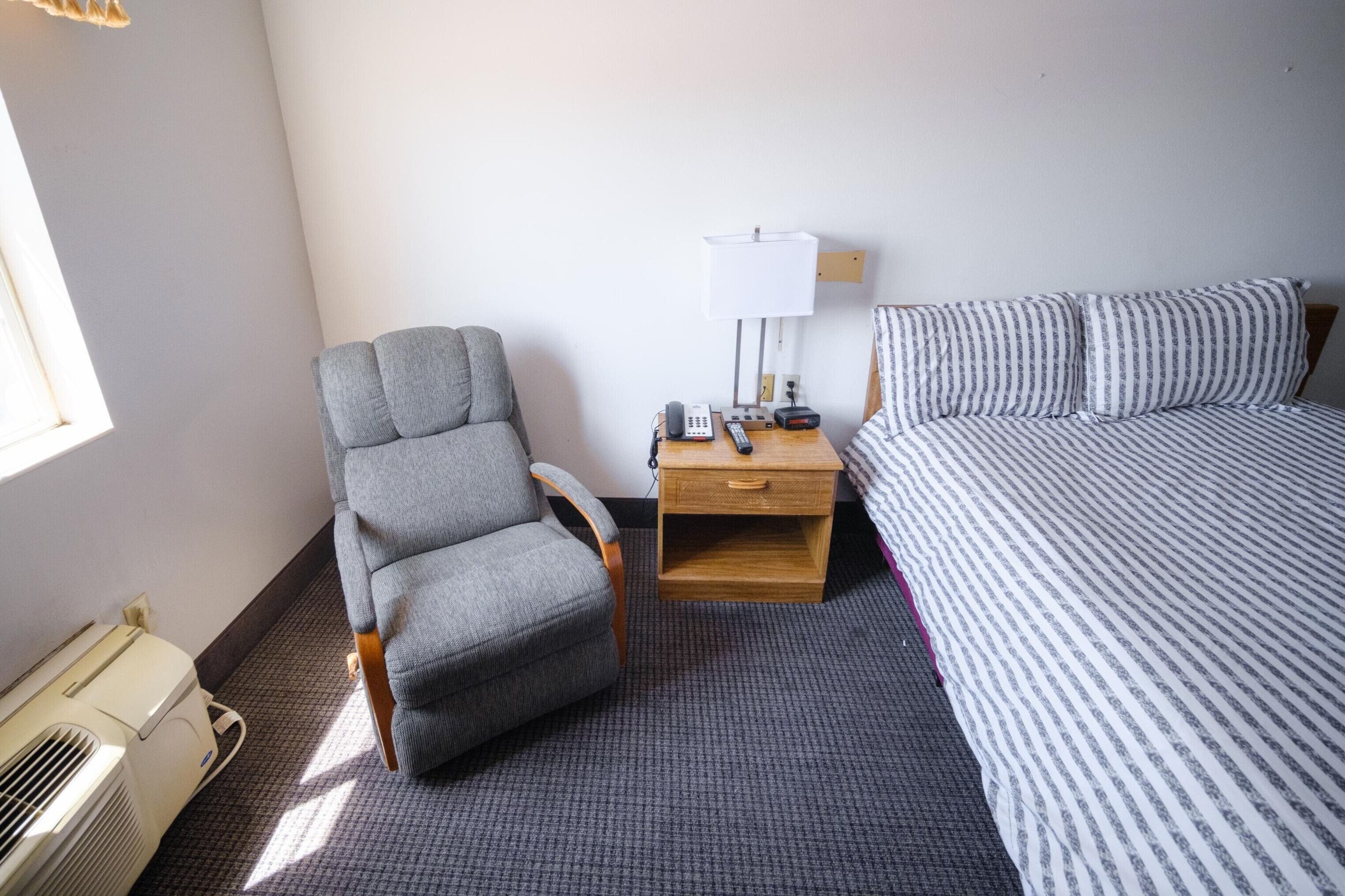 A hotel room with a single bed, a gray armchair, a wooden nightstand with a lamp, and a heater.
