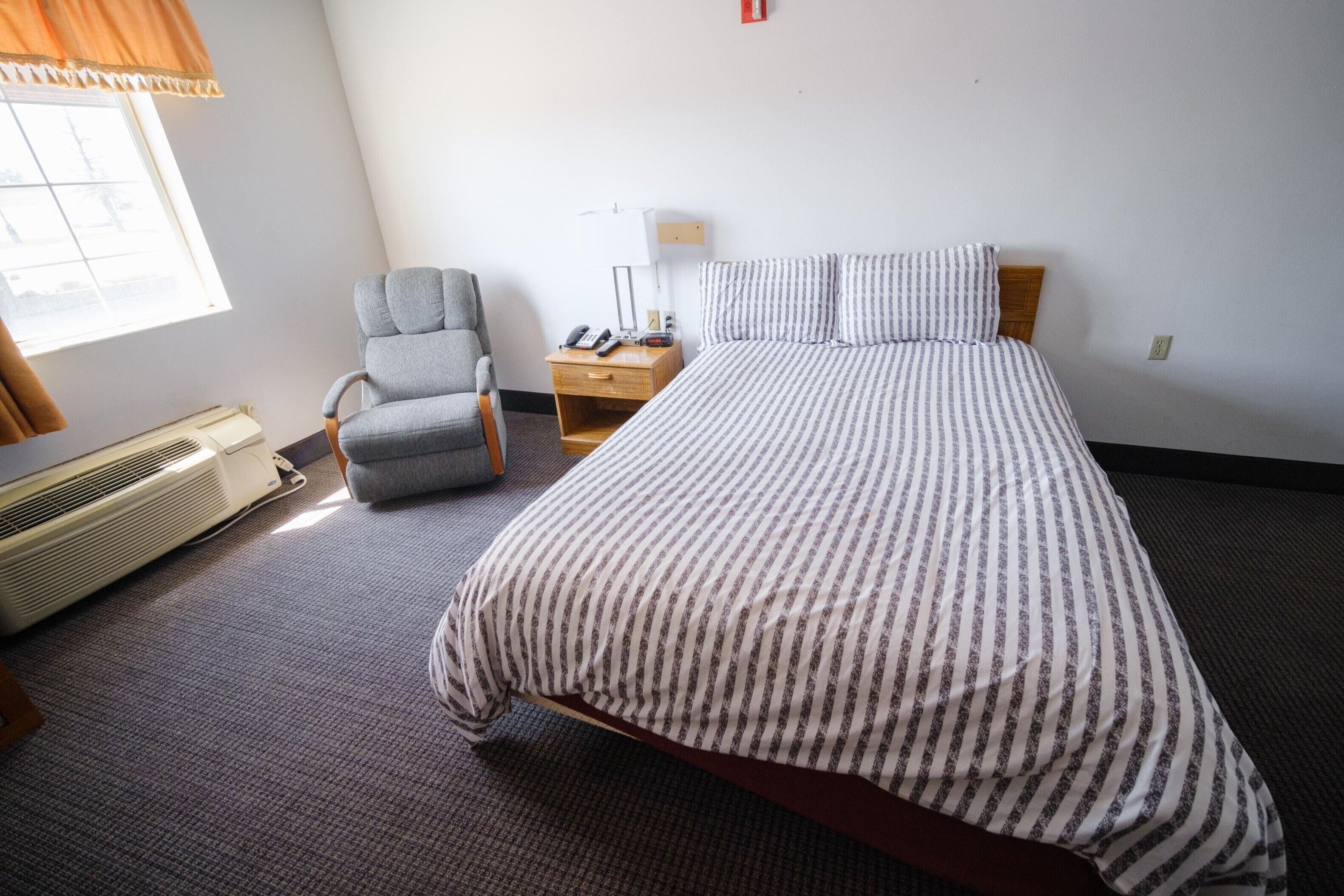 Una habitación de hotel con cama, una silla, una mesita de noche con lámpara y ventana.