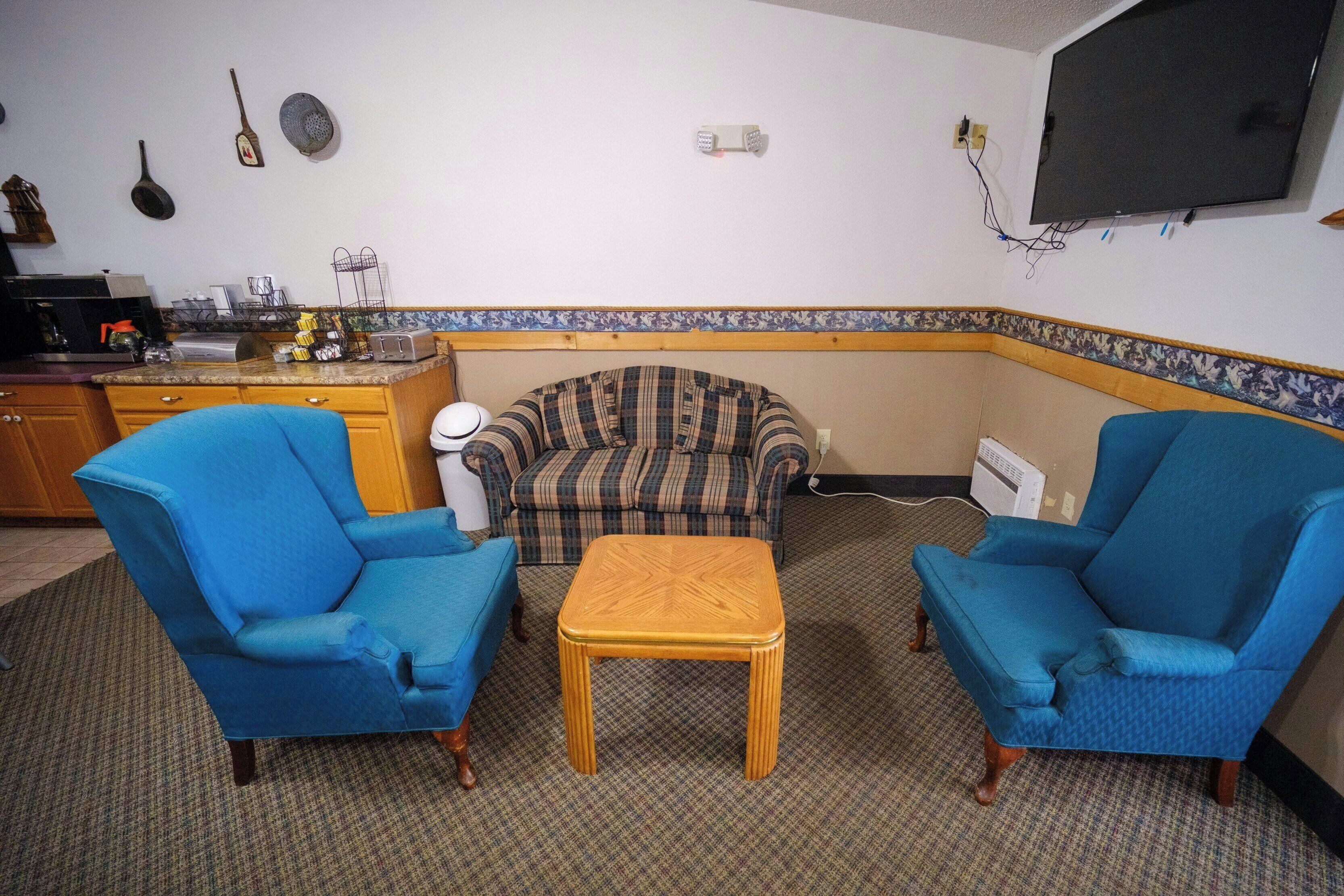 Uma sala de estar com dois poltronas azuis, uma mesa de centro de madeira, um sofá xadrez e uma área de cozinha com diversos objetos sobre o balcão.