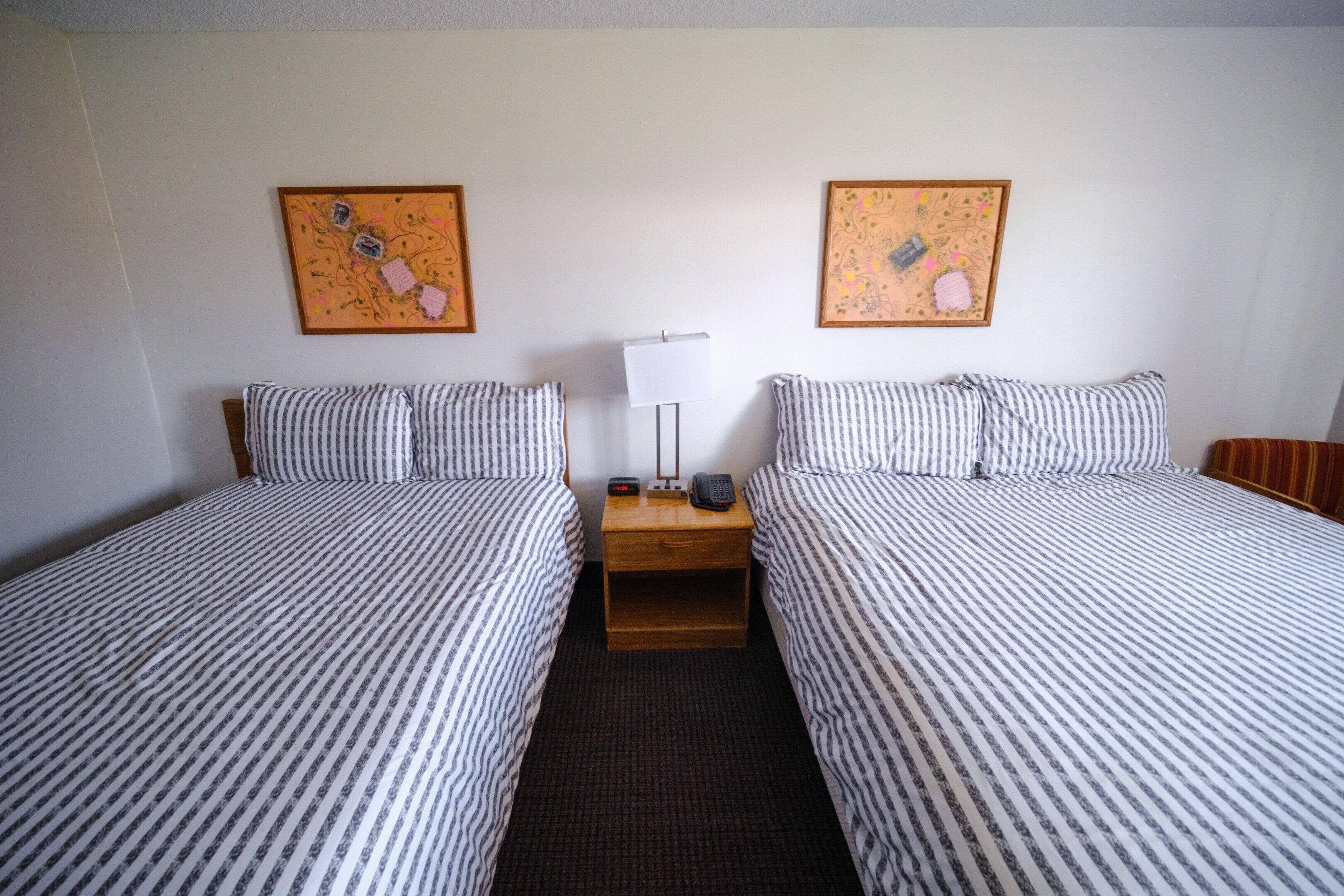 A hotel room with two beds, a nightstand with a lamp and phone, and two wall art pieces.