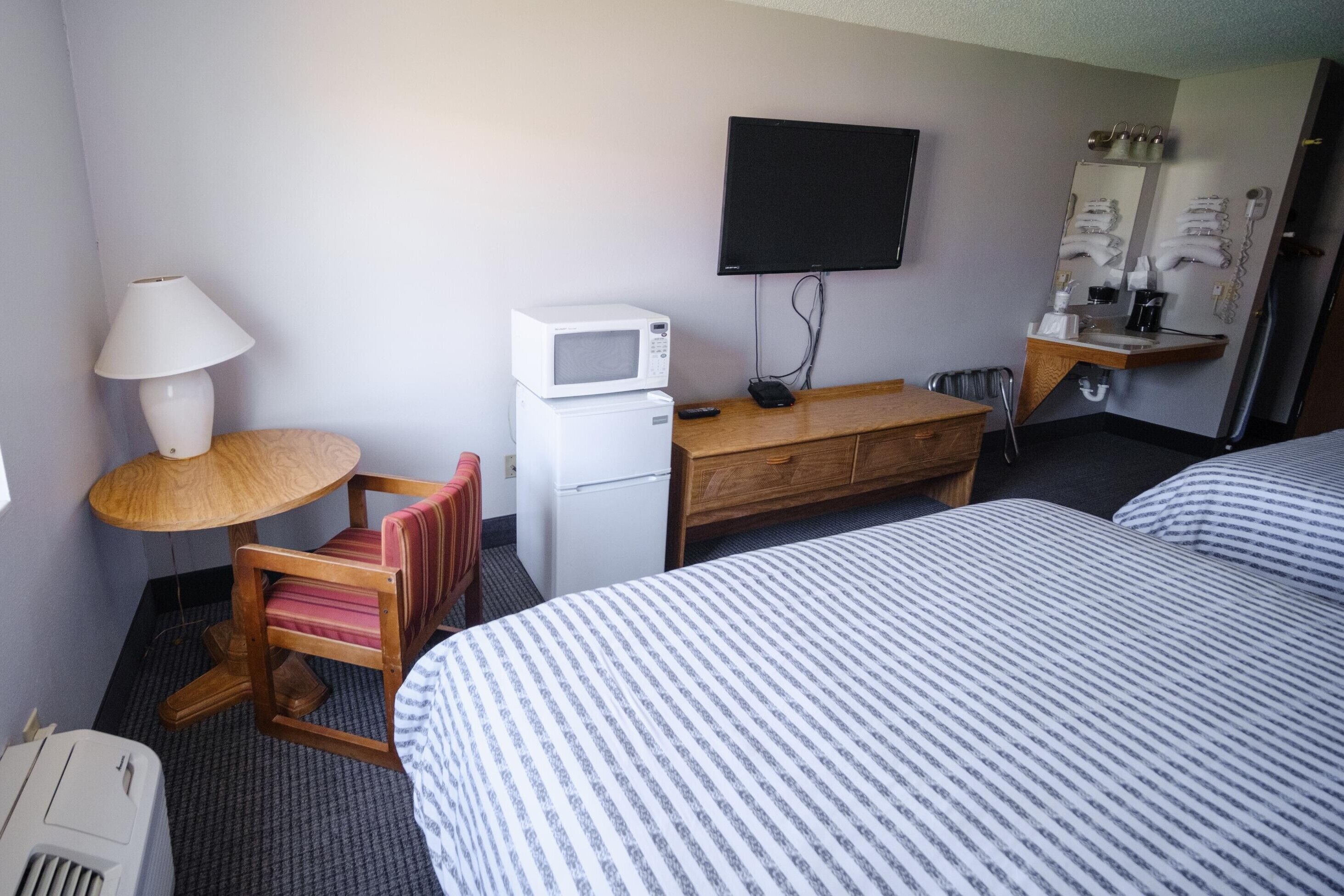 Habitación de hotel con una cama, un escritorio con microondas, una silla, un televisor y un tocador con espejo y artículos de aseo.