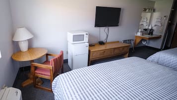 Habitación de hotel con una cama, un escritorio con microondas, una silla, un televisor y un tocador con espejo y artículos de aseo.