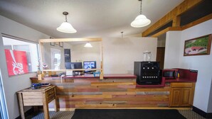 Une pièce avec un plancher en bois, un bureau avec un ordinateur, un four à micro-ondes et un réfrigérateur. Il y a une fenêtre avec une enseigne Coca-Cola, un miroir et un tableau accroché au mur.