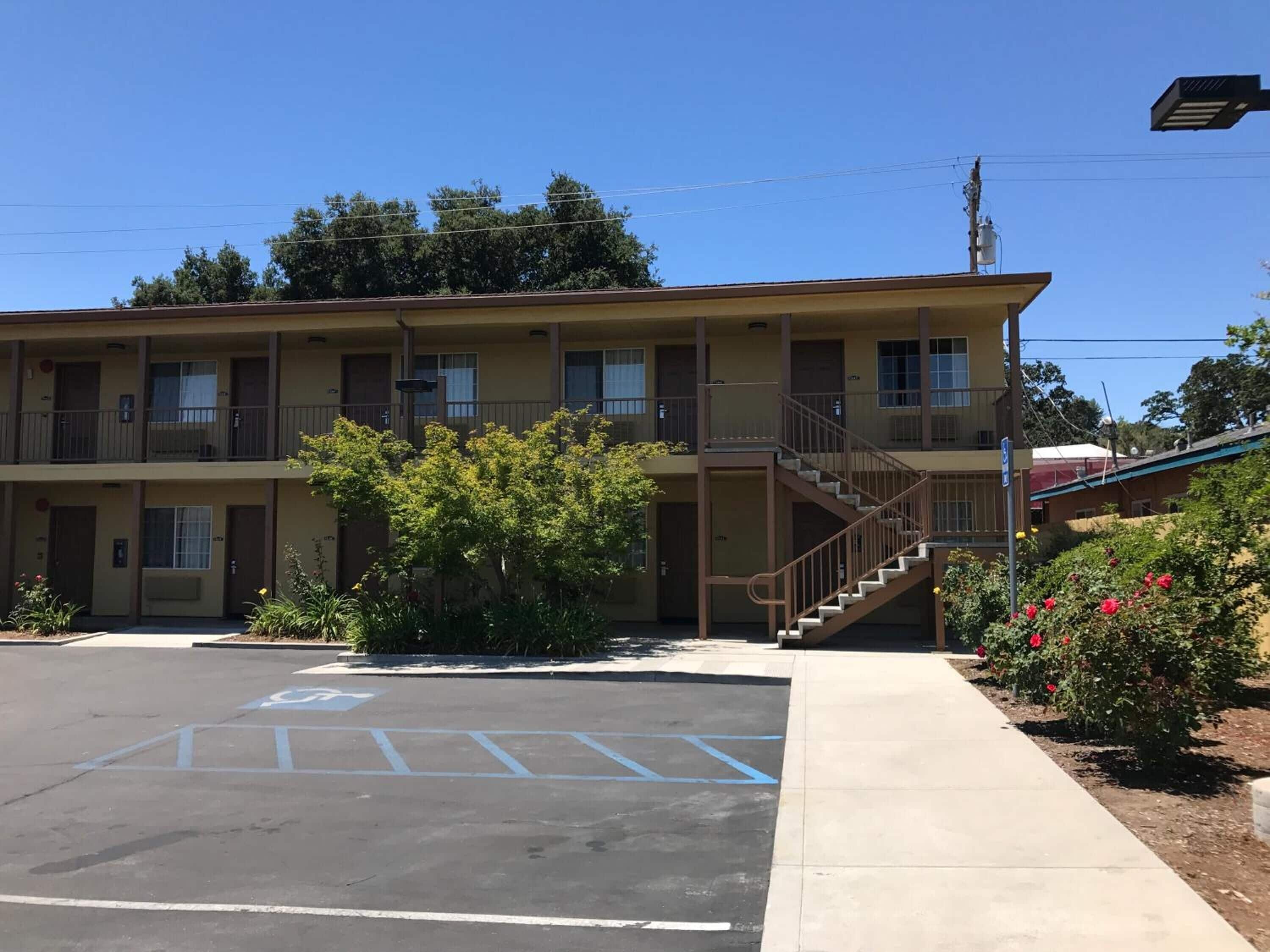 Un motel su due piani con una scala centrale, un parcheggio con posti riservati ai disabili e un piccolo giardino con fiori rossi.
