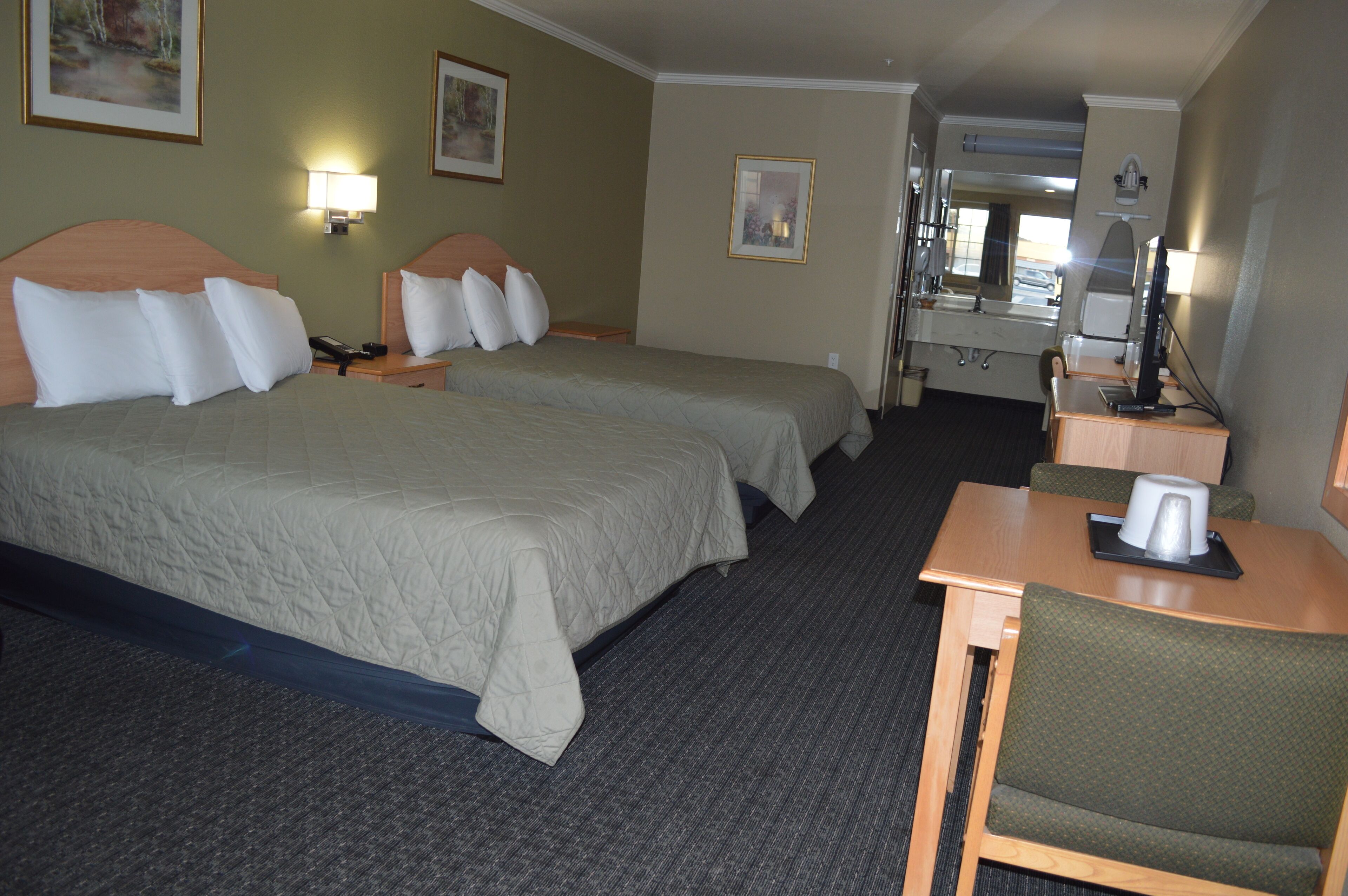 Quarto de hotel com duas camas, uma escrivaninha, uma cadeira, um espelho e um banheiro visível através de uma porta aberta.