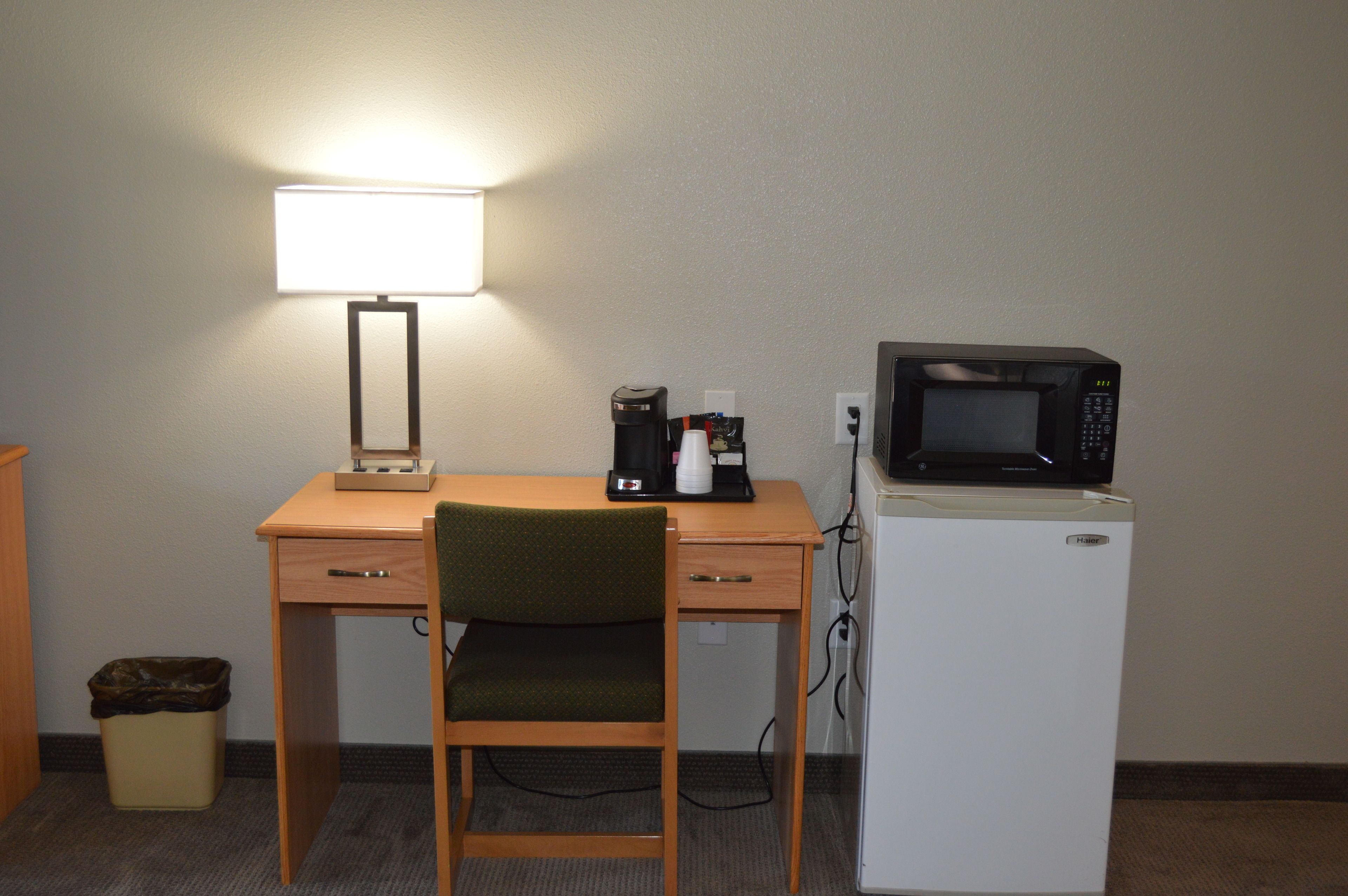 A hotel room with a desk, a chair, a microwave, a coffee maker, and a trash can.