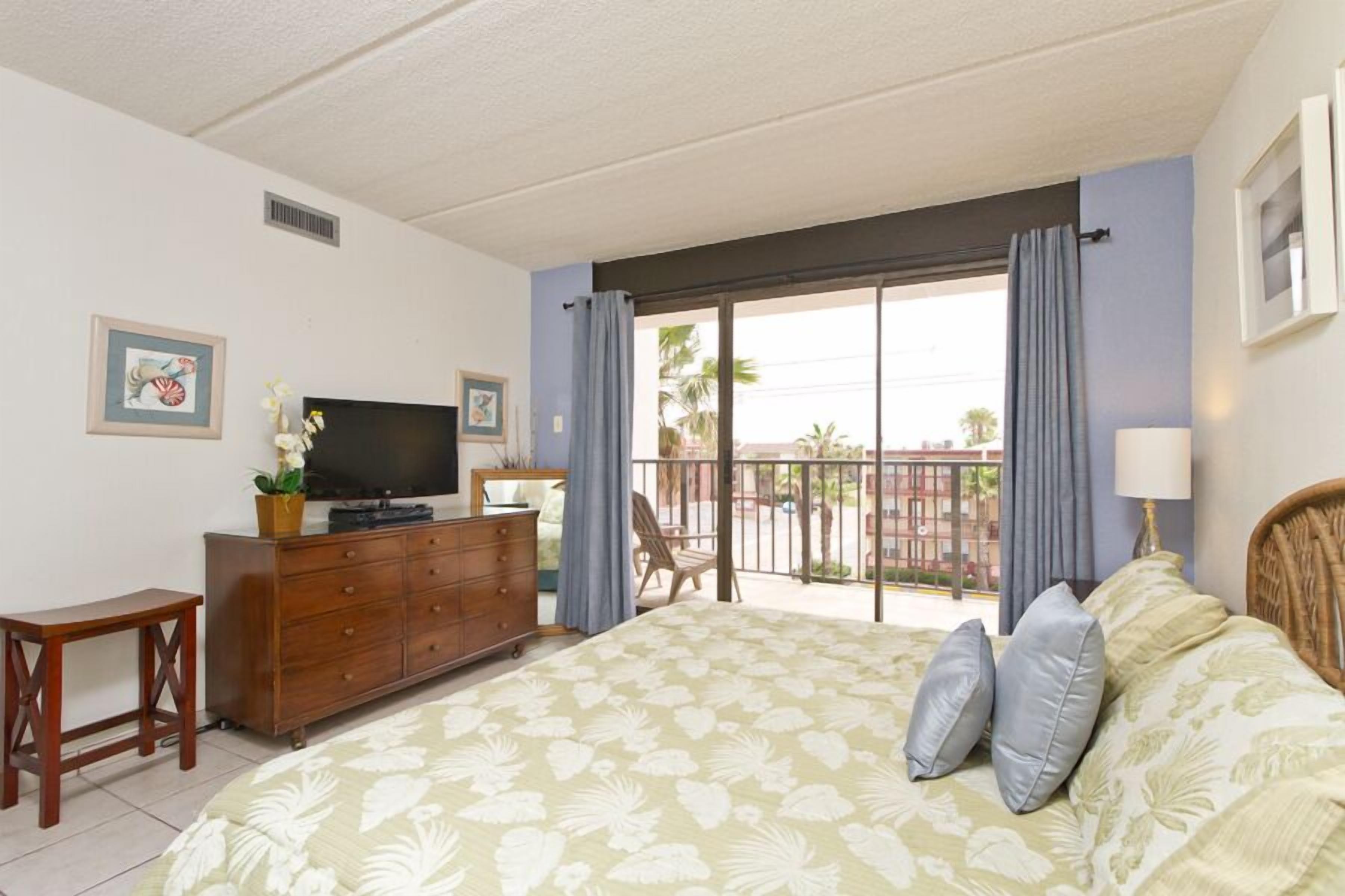 A bedroom with a bed, a dresser, a television, and a balcony with a view of palm trees.