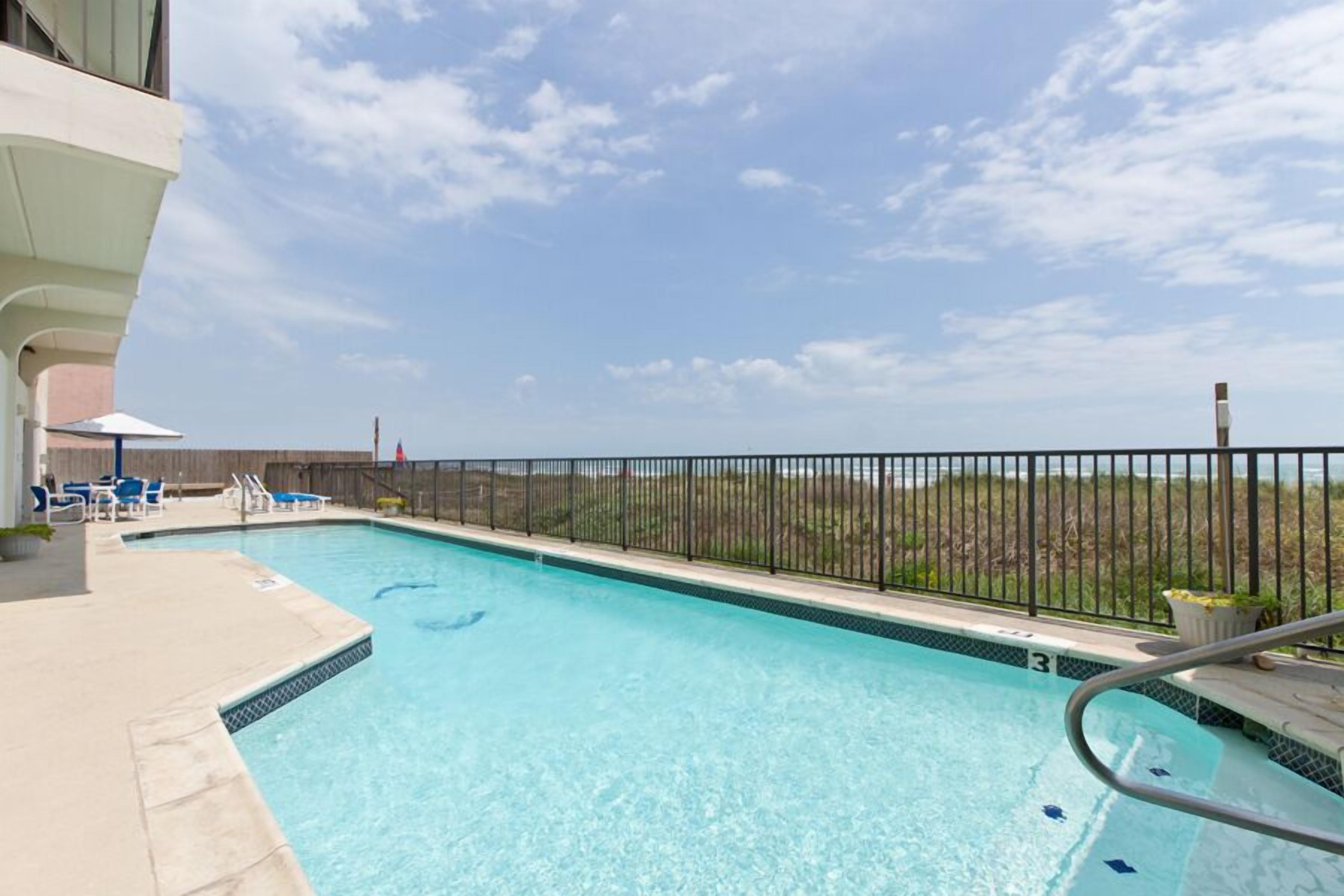 A swimming pool with a clear blue water, surrounded by a black metal fence, and a view of the beach in the background.