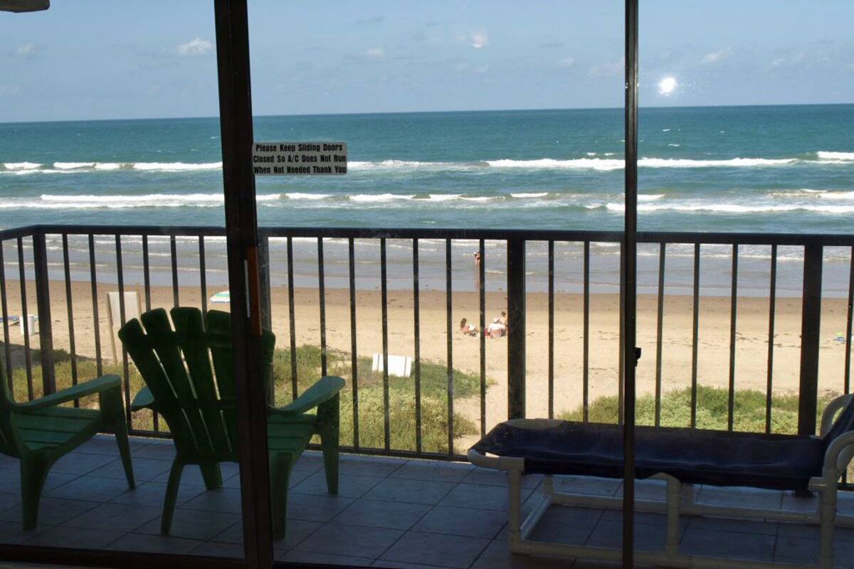 Un balcon avec vue sur la plage, un panneau indiquant de bien fermer les portes coulissantes, et des chaises vertes.