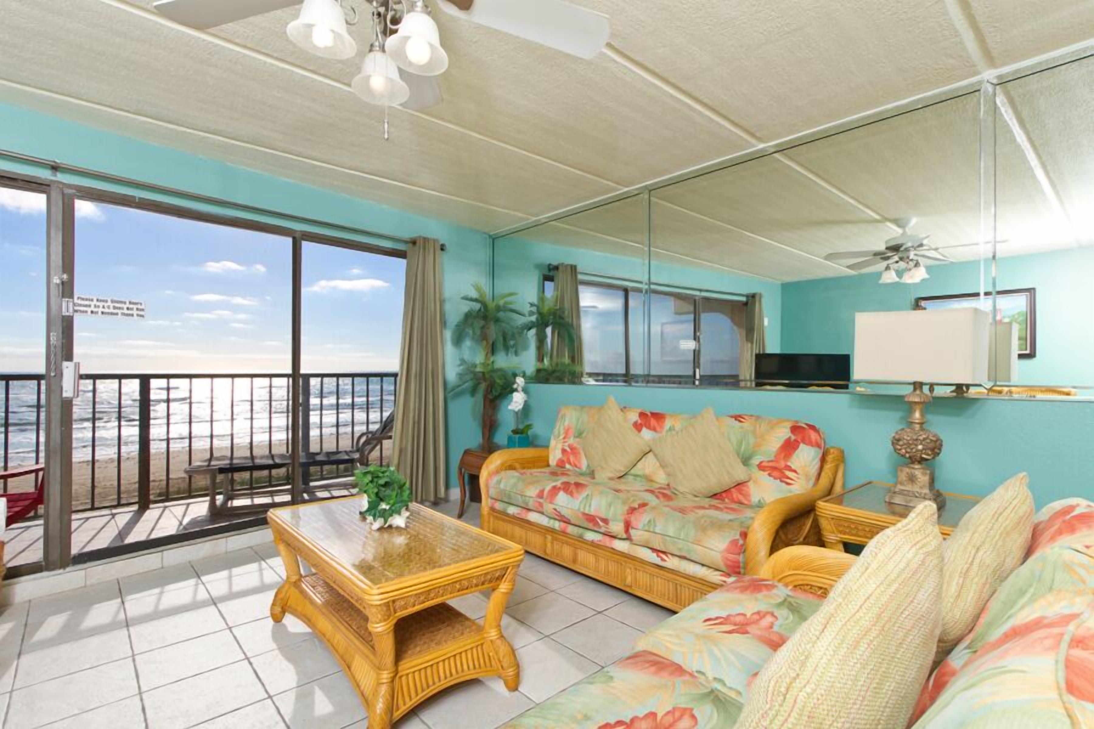 Uma sala de estar com sofá, mesa de centro e vista para a praia através de grandes janelas.