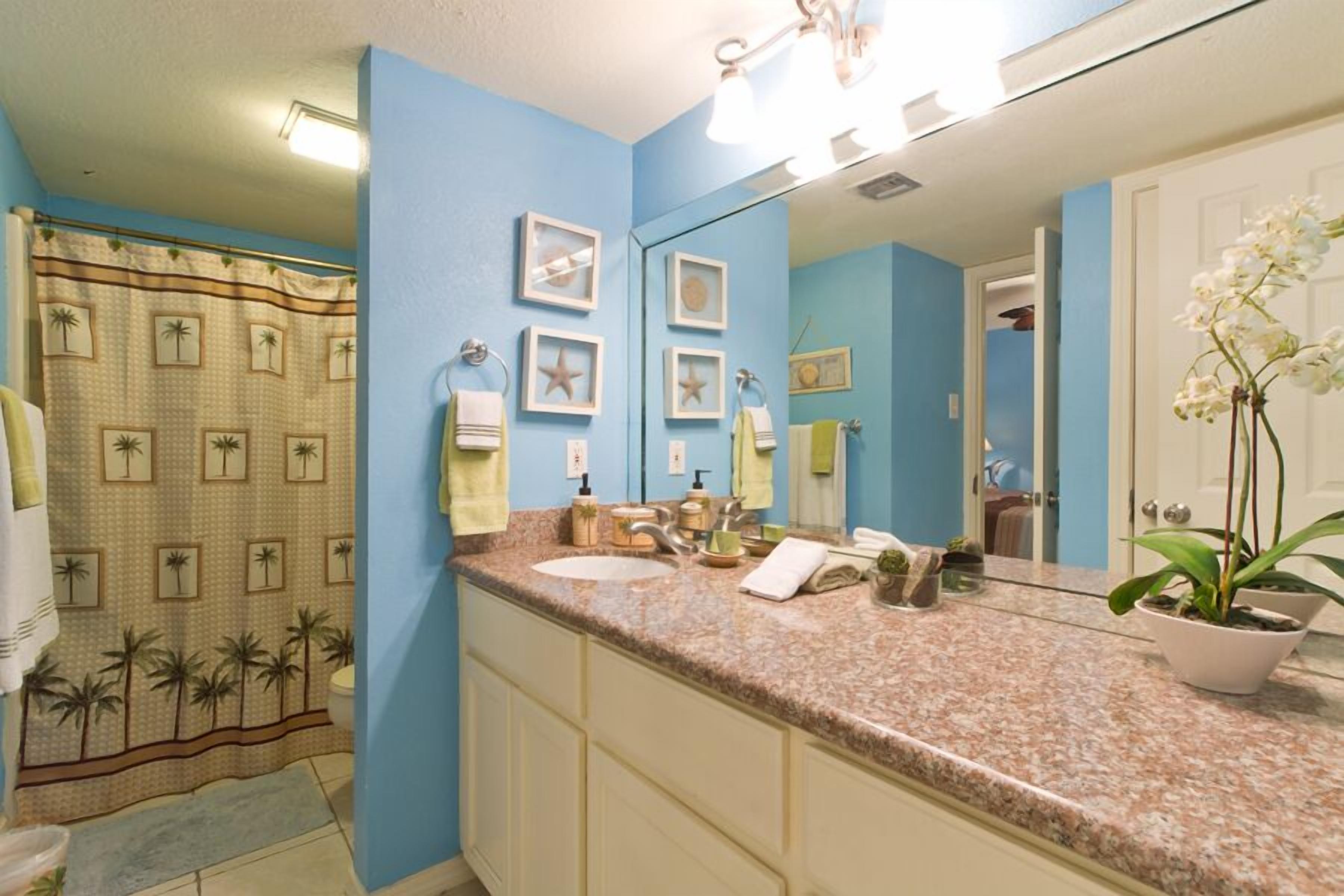 Bagno con parete blu, piano di lavoro in granito, lavabo, specchio e tenda da doccia con motivi tropicali.