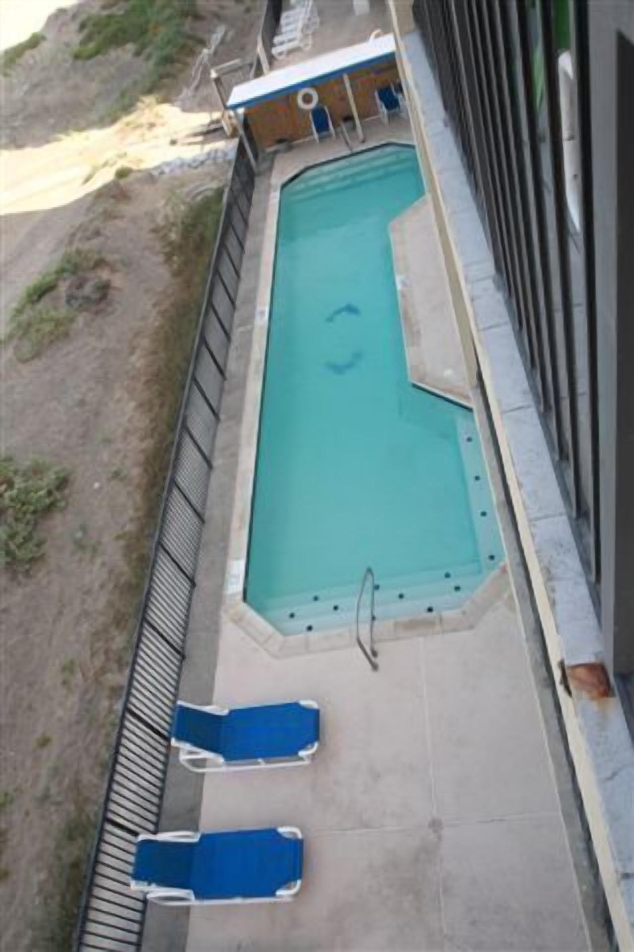 A rectangular swimming pool with a diving board, two blue lounge chairs, and a small building with a blue roof in the background.