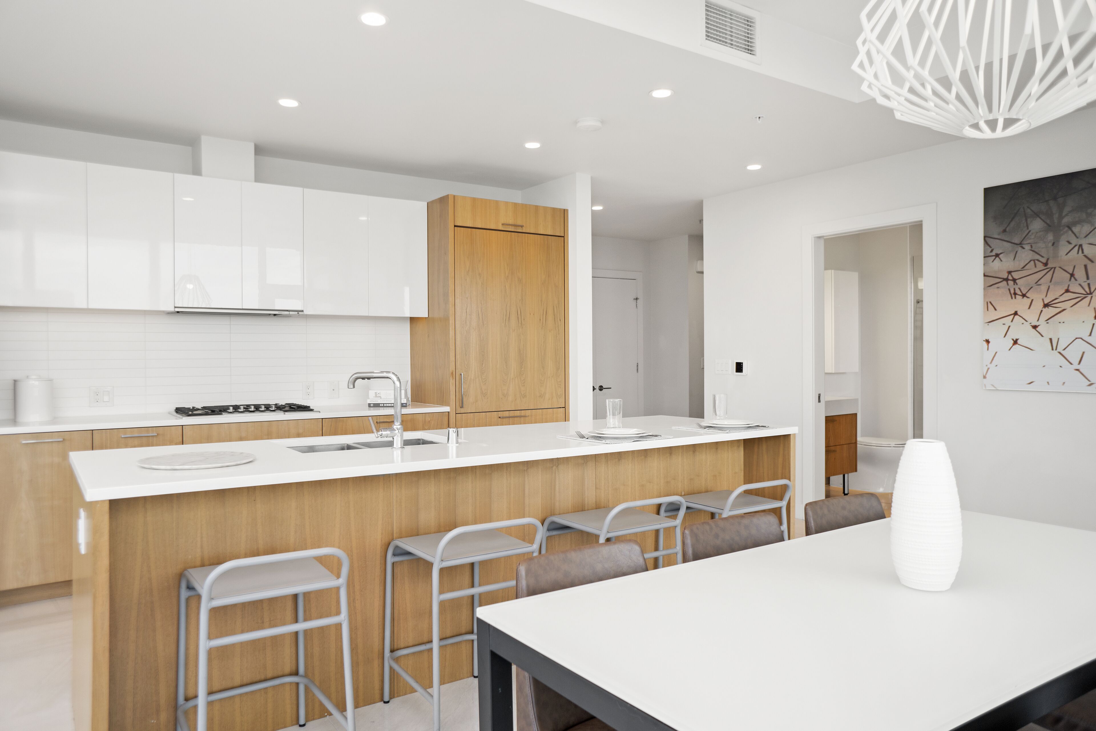 A modern kitchen with white cabinetry, a wooden accent wall, a central island with bar stools, and a white dining table with a vase.