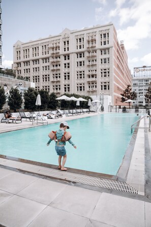 A swimming pool area with lounge chairs and umbrellas, a large building in the background, and a person with a backpack. - Level Los Angeles Downtown - South Olive (Los Angeles)