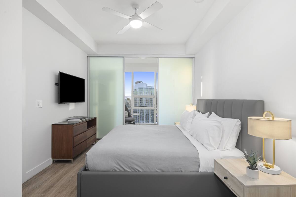 Een moderne hotelkamer met een bed, nachtkastje, lamp, televisie en uitzicht op een stadsbeeld door een schuifdeur.
