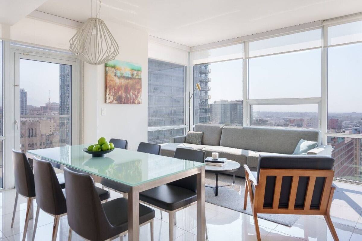 Een moderne woonkamer met een glazen eettafel, een grijze bank en grote ramen met uitzicht op de stad.