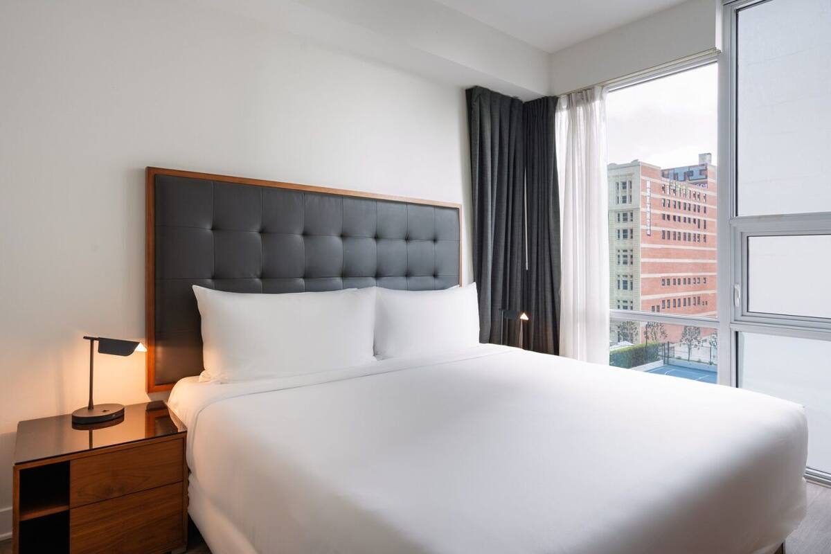 Een moderne hotelkamer met een groot bed, een houten nachtkastje, een lamp en uitzicht op gebouwen door een groot raam.