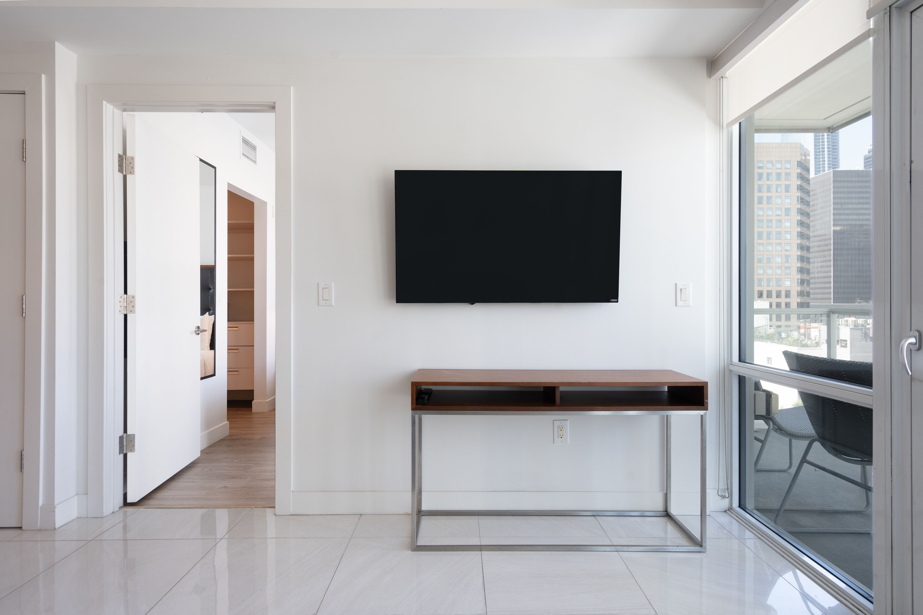 A modern room with a large window, a flat-screen TV mounted on the wall, and a wooden desk with metal legs.