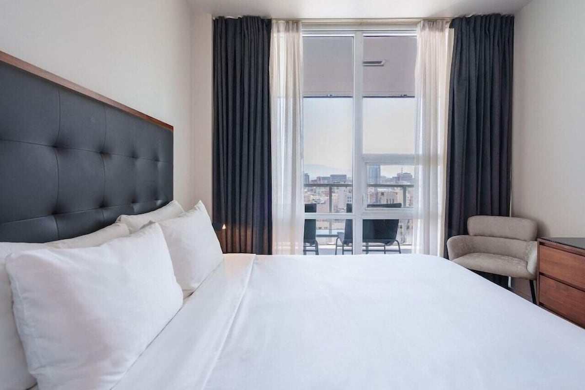 Hotellihuone, jossa on suuri sänky, tuoli, työpöytä ja ikkunasta avautuva näkymä kaupunkiin.