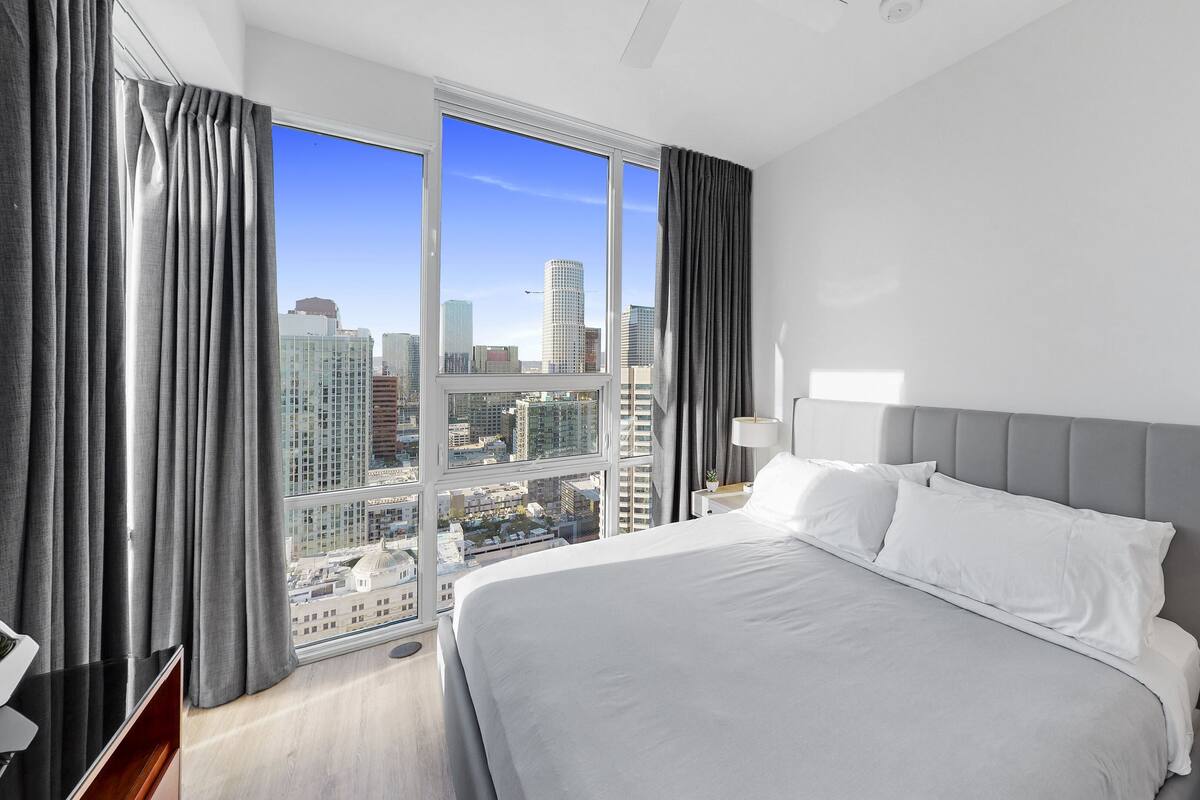 Een moderne hotelkamer met een groot bed, een nachtkastje met een lamp, en uitzicht op de stad door middel van ramen van de vloer tot het plafond.