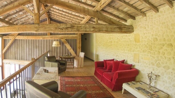 Un interior rústico con techo de madera, paredes de piedra, un sofá rojo, un sillón de cuero y una pequeña mesa con una lámpara.
