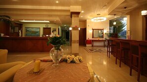 Un vestíbulo de hotel con una mesa de recepción, una mesa redonda con un florero y una vela, y una zona de bar con sillas de madera.