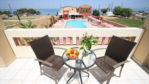 Un balcón con dos sillas de mimbre y una mesa de vidrio, con vista a una piscina y un edificio frente al mar.