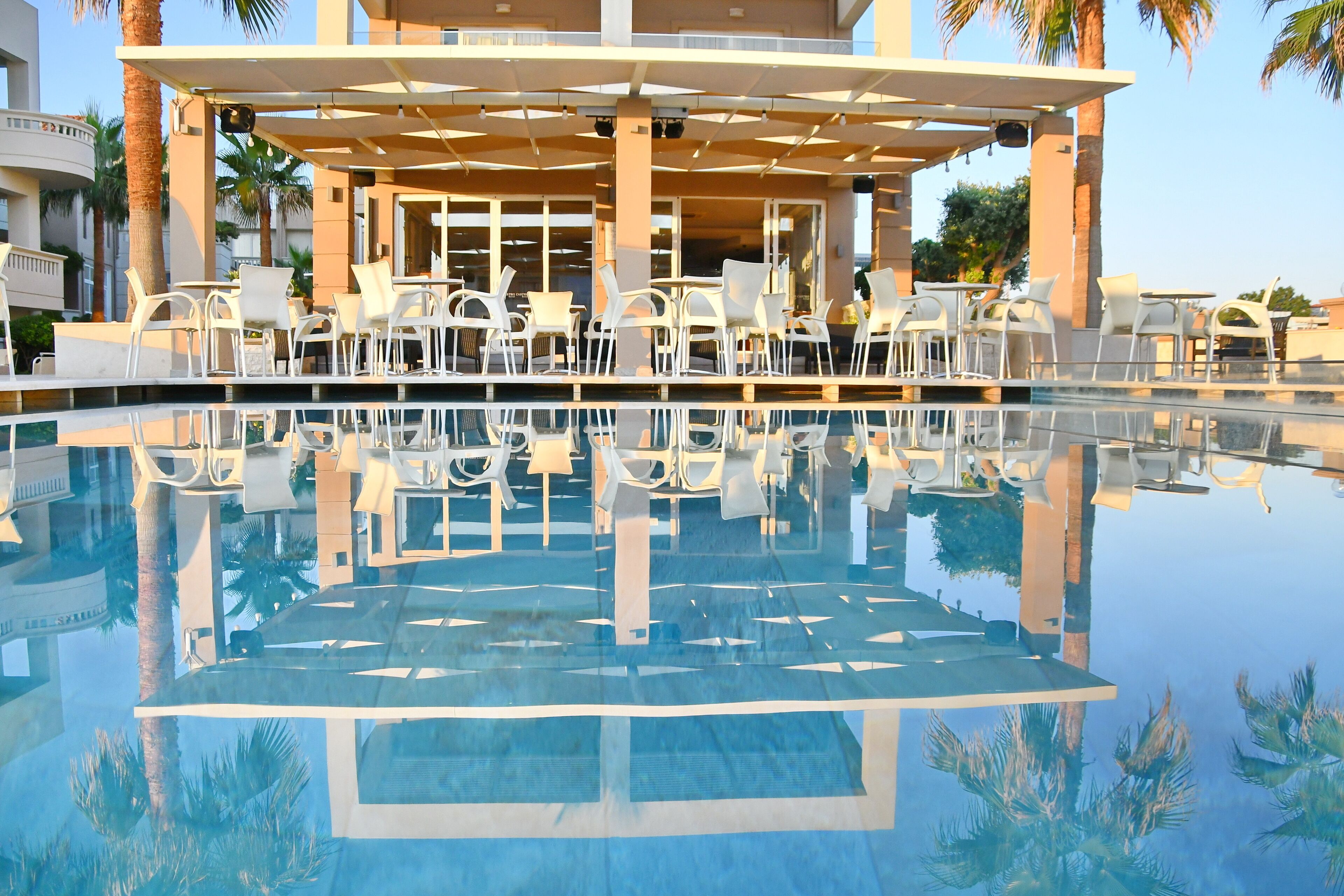 Uma área junto à piscina com cadeiras e mesas brancas, palmeiras e um edifício com janelas amplas.