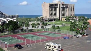 A large building with multiple floors, a parking lot with several vehicles, and a tennis court area.