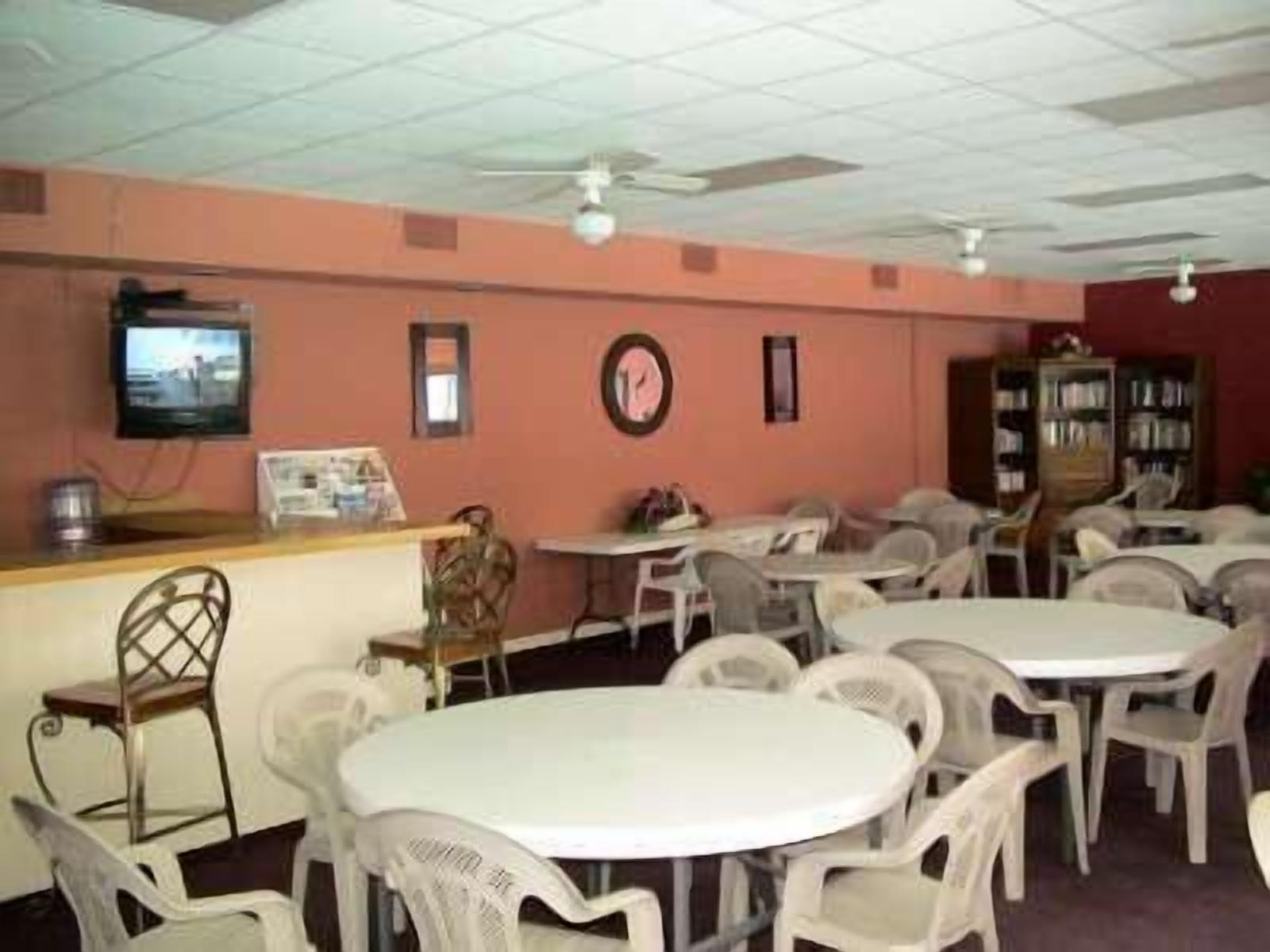 A room with round tables and plastic chairs, a television on the wall, and a bookshelf.