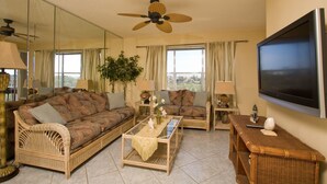 A living room with wicker furniture, a ceiling fan, a flat-screen TV, and large windows.