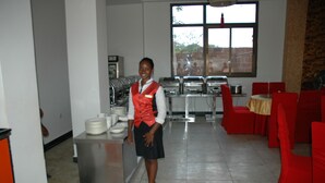 A hotel dining area with a buffet setup, a staff member, and red chairs. - Golden Chopstick Hotel (Livingstone)