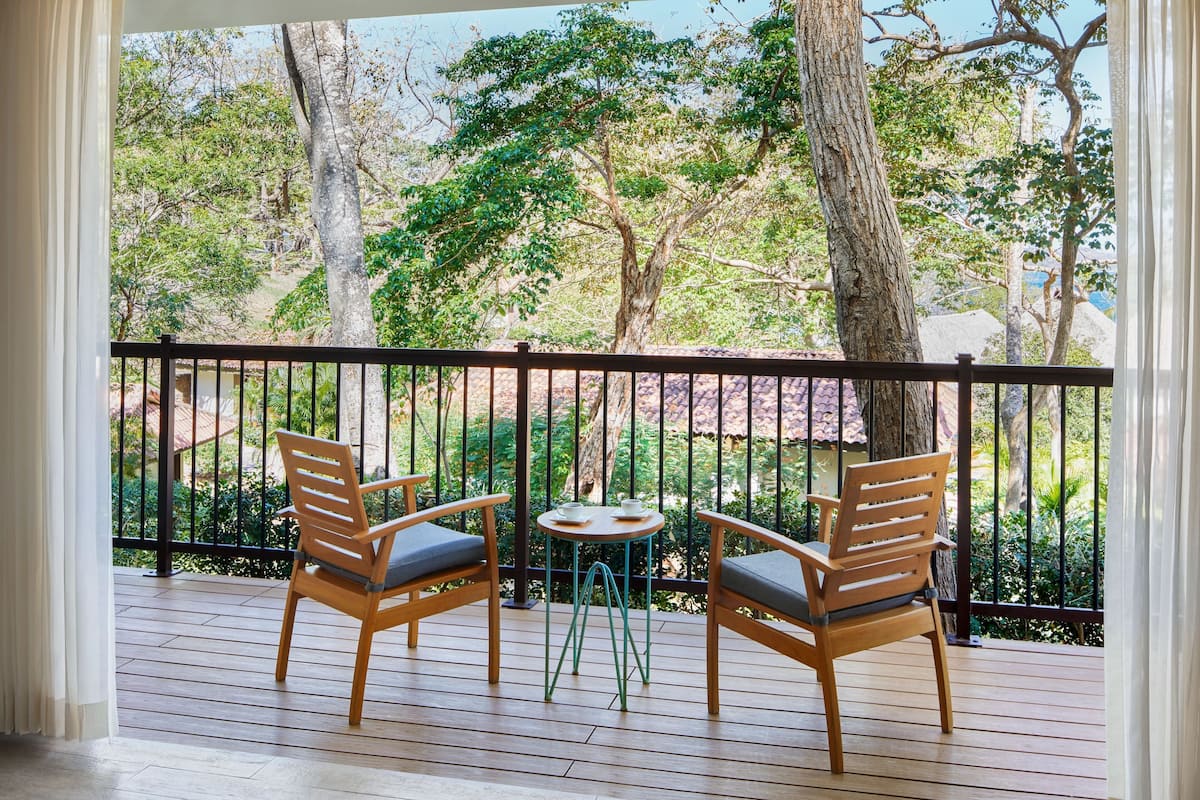 Une terrasse en bois, agrémentée de deux chaises et d’une petite table, entourée d’une rambarde noire, et offrant une vue sur un jardin luxuriant, avec des arbres et un bâtiment en arrière-plan.