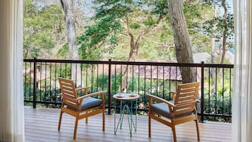A wooden deck with two chairs and a small table, surrounded by a black railing, overlooking a lush garden with trees and a building in the background.