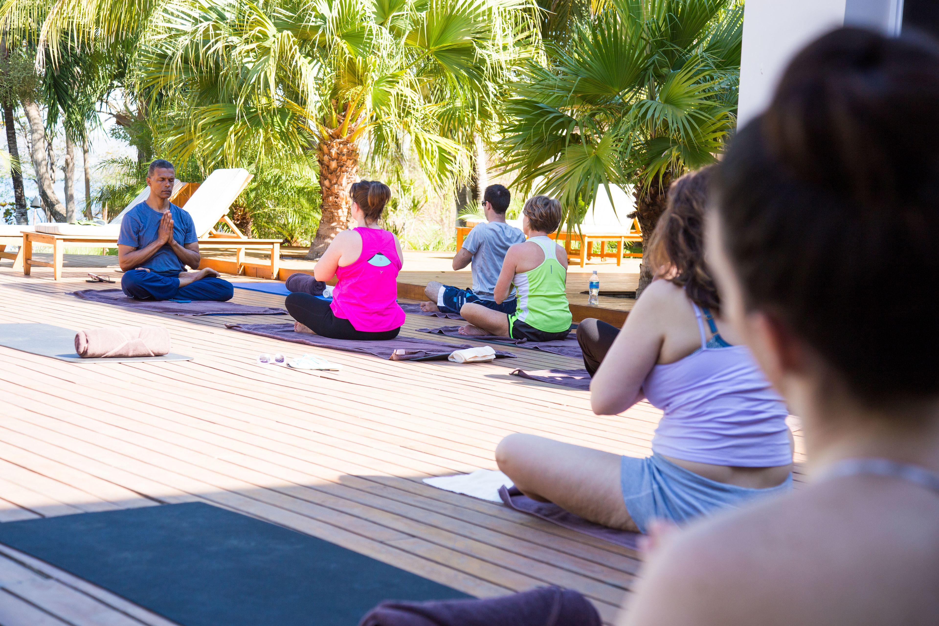 A group of people sitting on yoga mats outdoors on a wooden deck, surrounded by palm trees.