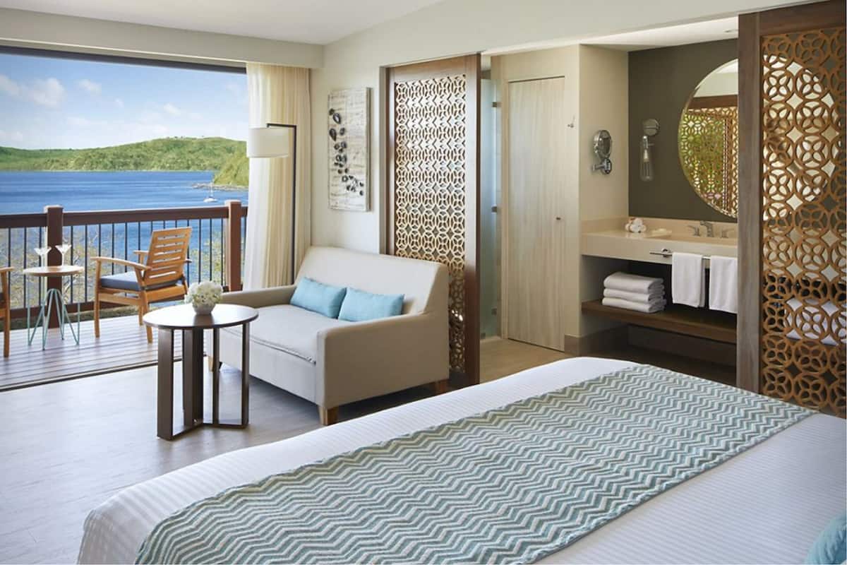 Une chambre d’hôtel avec un balcon, un canapé, un lit, une petite table et une salle de bain équipée d’un miroir et d’un porte-serviettes.