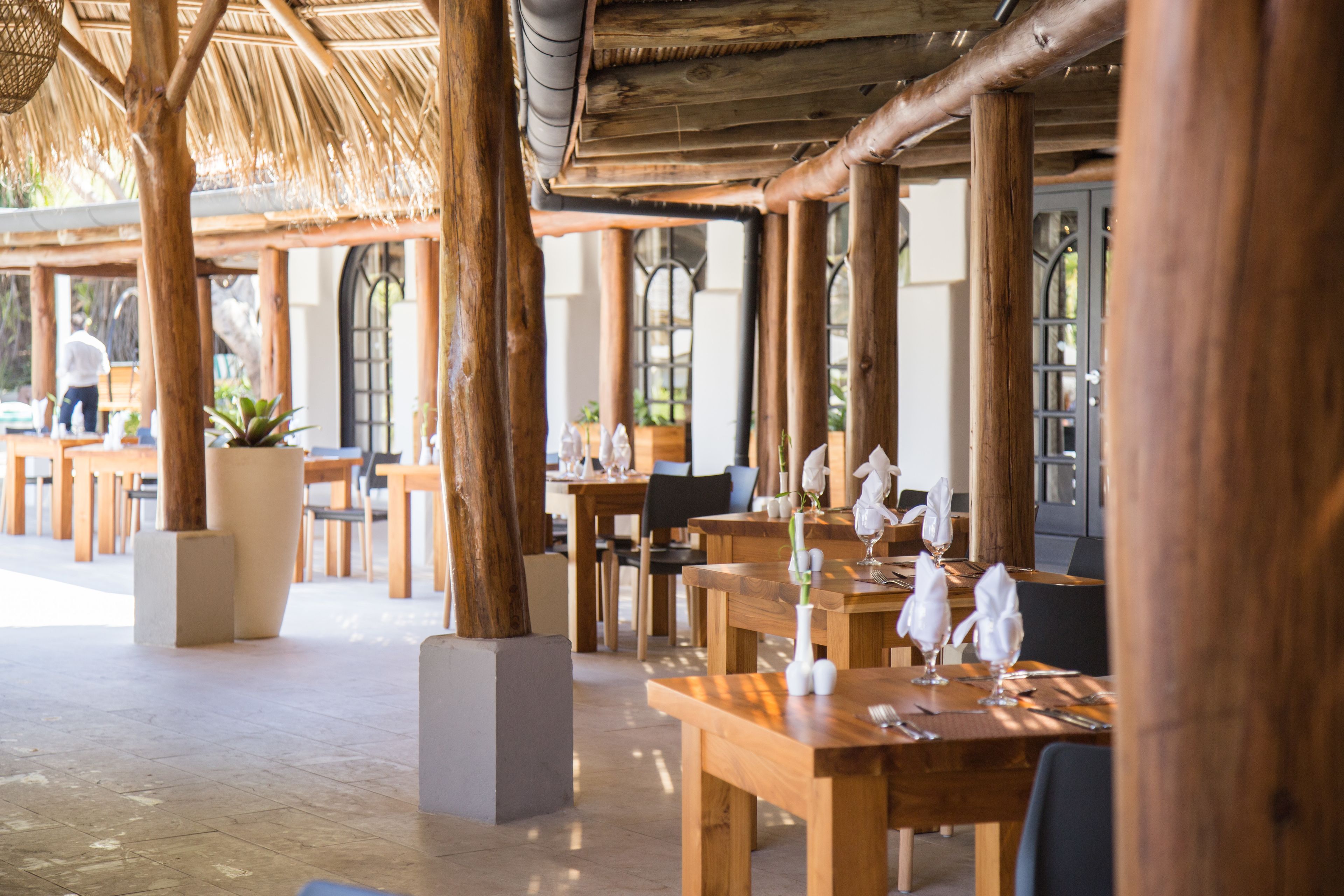 An outdoor dining area with wooden tables and chairs, large potted plants, and a thatched roof structure.
