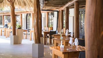 An outdoor dining area with wooden tables and chairs, large potted plants, and a thatched roof structure.