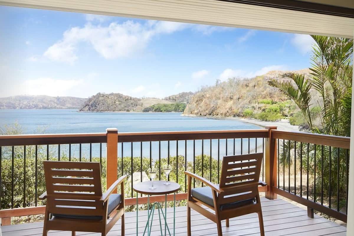 Une terrasse en bois avec deux chaises et une petite table, offrant une vue sur un lac et un paysage vallonné.