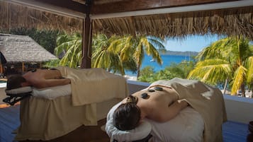 Relax in a serene spa setting overlooking the ocean, with two massage tables under a thatched roof.