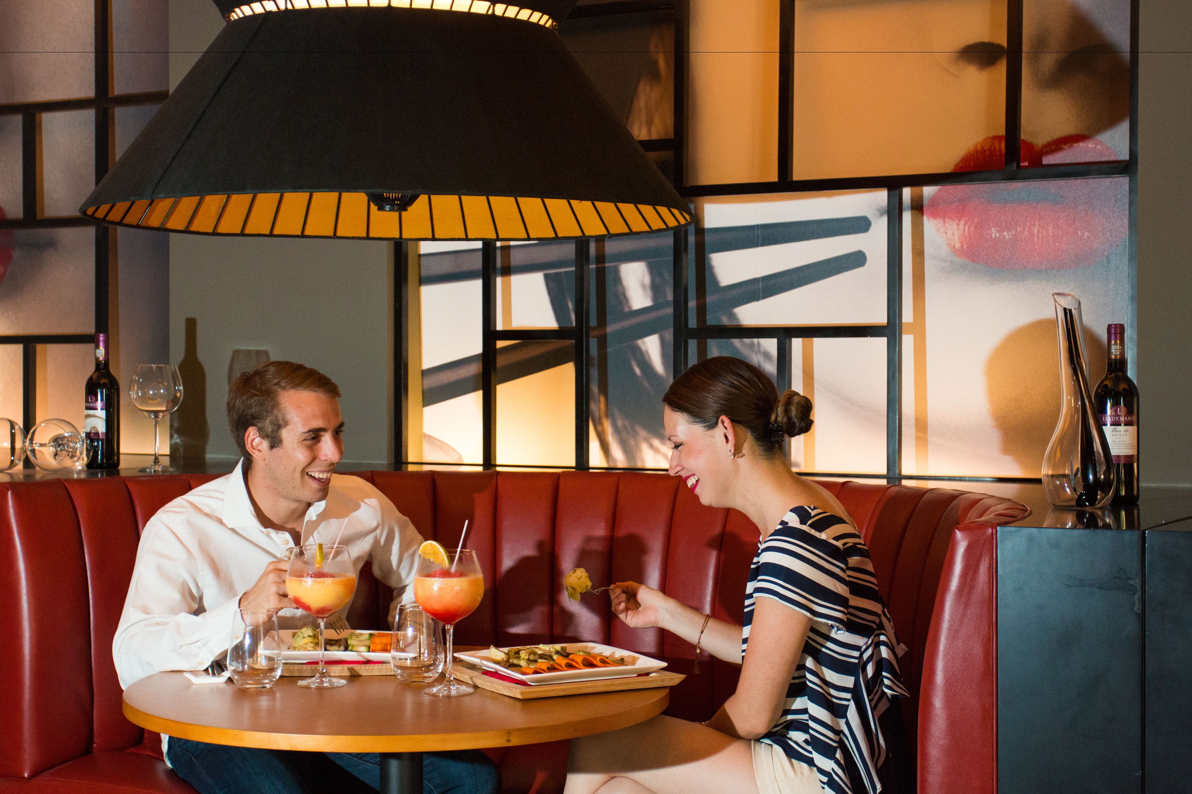 A couple dining at a restaurant with a red booth, a round table, and a large black lamp overhead.