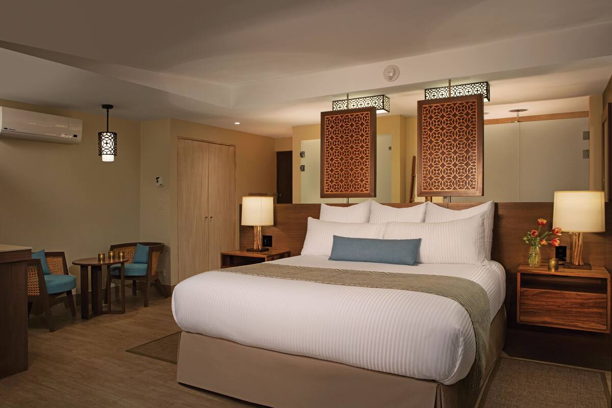 Une chambre d’hôtel avec un grand lit, deux tables de chevet avec des lampes, une petite table avec des chaises et un élément de séparation décoratif.