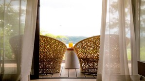 Une pièce avec deux fauteuils en osier placés face à une table sur laquelle se trouve un verre de jus d’orange, et une vue sur la verdure et les montagnes, visible à travers de fines rideaux.
